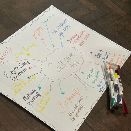 A sheet of papaer on the ground with a pack of felt tips open next to it. On the paper in different colours saying Self Care Tips